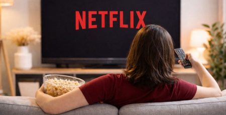 Mulher jovem sentada no sofá com controle de tv e pipoca na mão com tela de TV na frente para assistir lançamentos netflix 2026